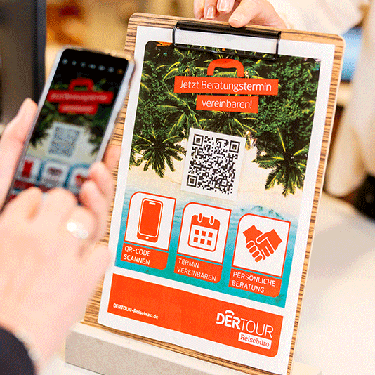 Ein Foto zeigt einen Aufsteller von DERTouristik, womit man sich per QR-Code einen Beratungstermin buchen kann. Im Vordergrund hält eine Hand ein Smartphone auf den QR-Code.