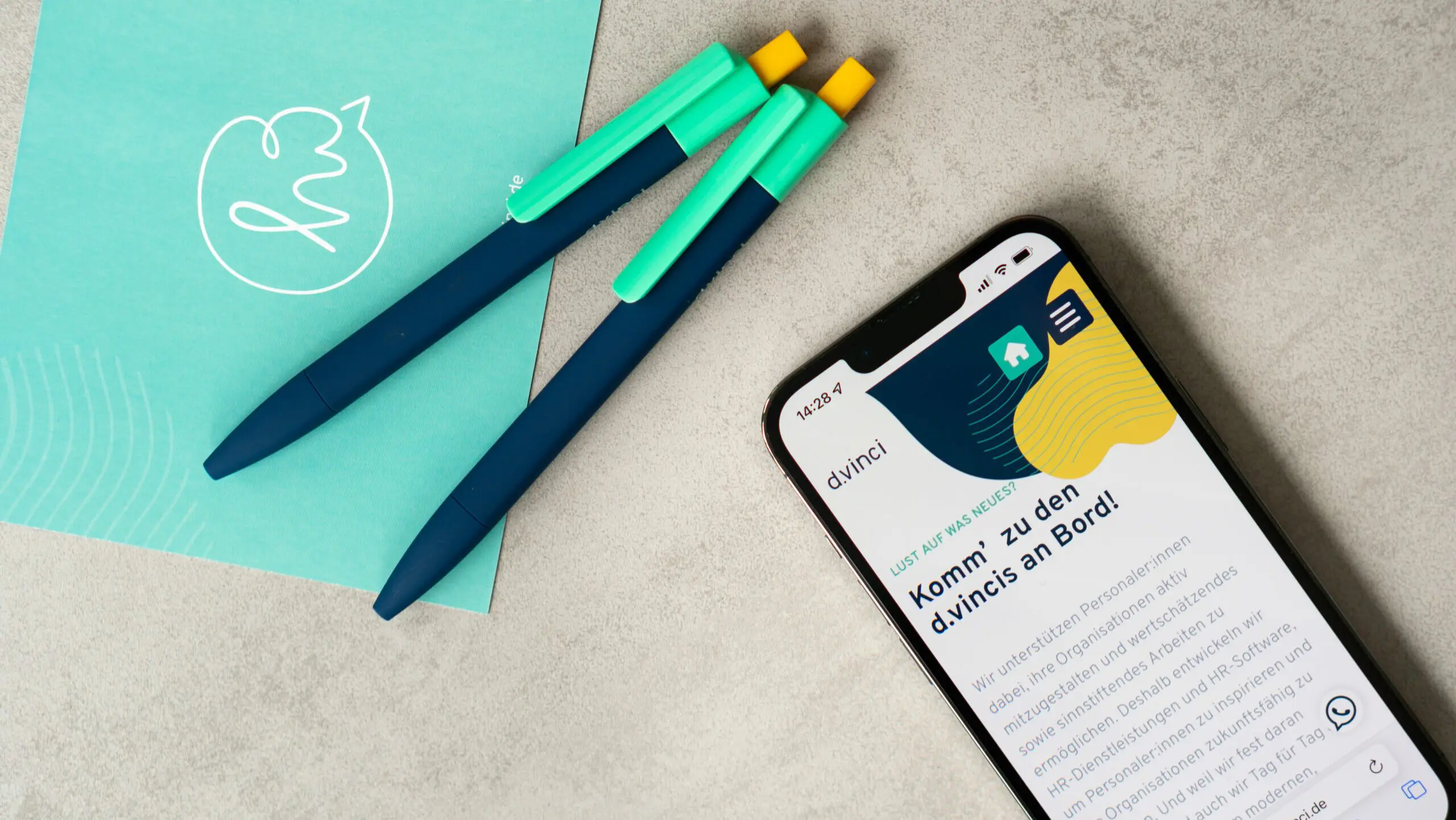Ein Foto zeigt ein Smartphone mit einem Ausschnitt der d.vinci-Karriereseite. Darüber liegen auf grauem Untergrund d.vinci-Kugelschreiber in türkis und dunkelbau und eine türkise d.vinci-Postkarte mit einem Hi-Icon.